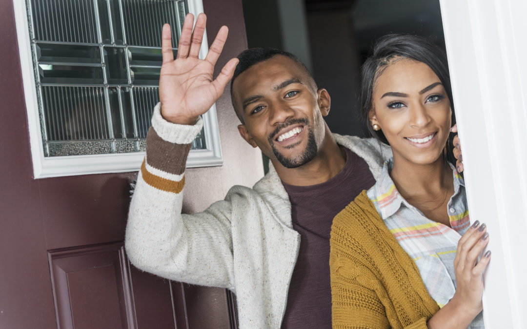 A young couple smiling, standing together in the front doorway of their home. The woman is mixed race black and Caucasian and the man is African-American. They received a welcome packet when they moved into their community association.