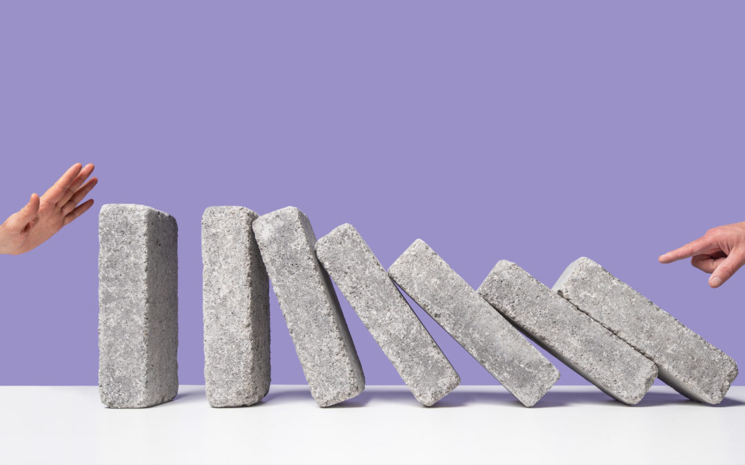 One man's hand starting domino effect with concrete blocks, another man's hand poised to stop blocks from falling at opposite side, purple background, white surface
