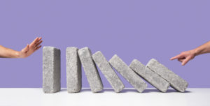 One man's hand starting domino effect with concrete blocks, another man's hand poised to stop blocks from falling at opposite side, purple background, white surface
