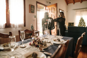 Coming over for Christmas dinner during the COVID-19 pandemic. Two women greet each other at the entrance of a house, with a Christmas decorated table for dinner