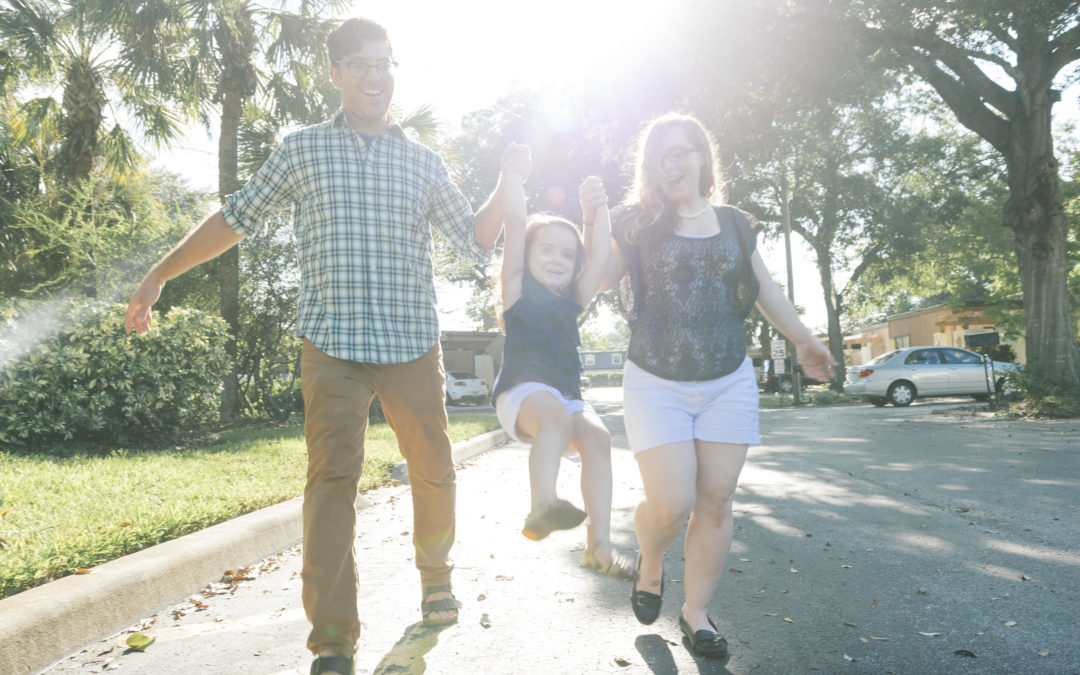 This is a horizontal, color photograph of a young American family spending time outdoors on a sunny day in their Winter Park neighborhood, a suburb of Orlando, Florida. A couple walks holding hands with a 4 year old girl who is swinging between them as they take a walk. The bright sun shines down through the trees.